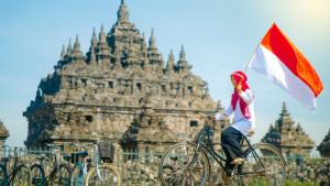 Indonesia tourism ranking in 2024 illustrated with a woman riding a bicycle and holding the Indonesian flag in front of a historic temple.