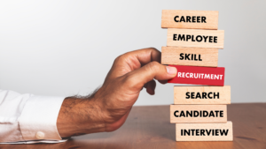 Recruitment Trends in 2025 : A close-up of a hand pulling a red block labeled 'Recruitment' from a stack of wooden blocks with words such as 'Career,' 'Skill,' and 'Employee,' representing the key elements of hiring strategies.