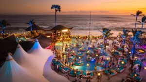 A lively Bali Beach Club at sunset with guests enjoying the pool, ocean views, and vibrant lighting along the beachfront.