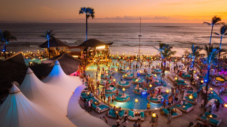 A lively Bali Beach Club at sunset with guests enjoying the pool, ocean views, and vibrant lighting along the beachfront.