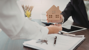 Two people exchanging a house model over property documents during a legal contract process to transfer property ownership in Indonesia.